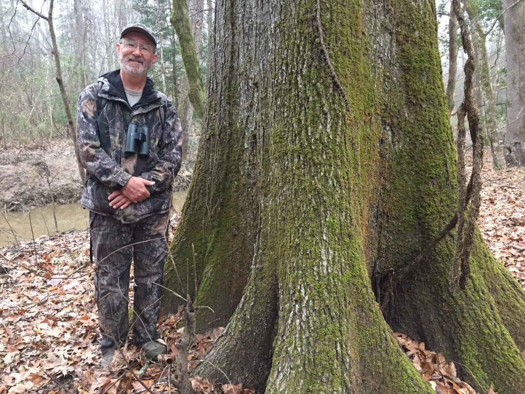 John Trochet wearing camouflage standing next to a large tree in the woods