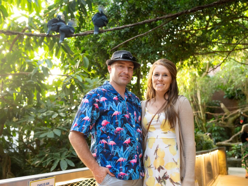 A husband and wife posing in the Tropical Rainforest with three Victoria Crowned Pigeons perched on a branch behind them.